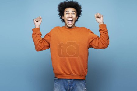Téléchargez les photos : Heureux jeune homme afro-américain tenant la main vers le haut, criant, succès de célébration isolé sur fond bleu. Concept gagnant - en image libre de droit