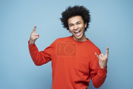 Foto de Young emotional African American man screaming, showing rock sign isolated on blue background - Imagen libre de derechos