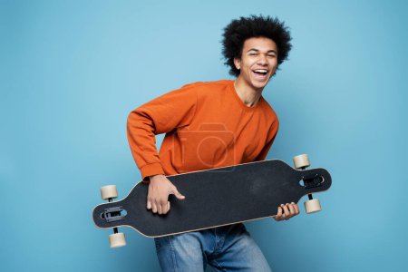 Foto de Happy attractive African American man holding longboard simulate playing guitar, having fun isolated on blue background. Portrait of attractive overjoyed skater looking at camera. Positive emotions - Imagen libre de derechos