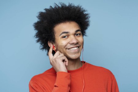 Téléchargez les photos : Authentique portrait de l'homme afro-américain souriant attrayant écoutant de la musique dans des écouteurs rouges regardant la caméra isolée sur fond coloré. Concept des personnes et des émotions - en image libre de droit