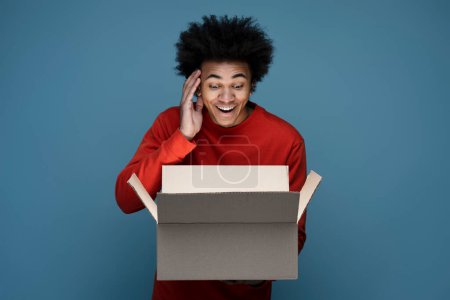 Photo for Young happy African American man influencer unpacking delivery box isolated on blue background. Excited customer opening package with birthday gift. Online shopping, sale concept - Royalty Free Image