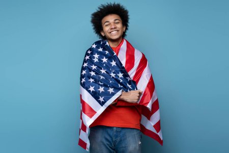 Téléchargez les photos : Confident smiling African American man covering with American flag isolated on blue background. Young emotional patriot celebration Independence day looking at camera. July 4th - en image libre de droit