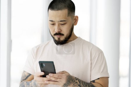 Foto de Serious guapo asiático hombre usando el teléfono móvil, compras en línea en casa. Hipster chico con elegante tatuaje de chat, la comunicación en línea. Concepto tecnológico - Imagen libre de derechos