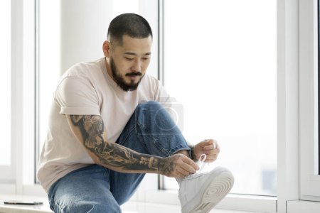Photo for Pensive handsome asian man with stylish tattoos tying shoelaces sitting on the windowsill. Young Korean guy wearing casual clothing at home - Royalty Free Image