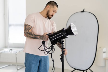 Foto de Professional Asian photographer adjusts lamp with lighting, preparing for shooting.  A young man works in a professional photo studio - Imagen libre de derechos