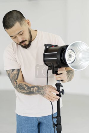 Photo for Portrait of professional asian photographer with stylish tattoos setting up lighting in modern studio. Assistant preparing for photo shoot - Royalty Free Image