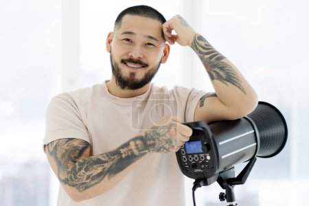 Portrait of smiling asian photographer setting up his lighting looking at camera in photo studio