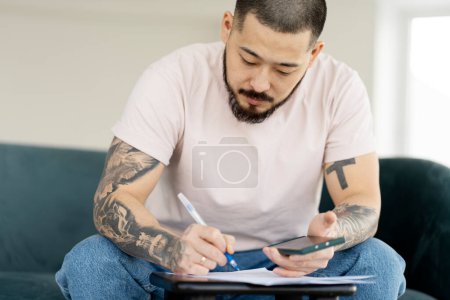 Téléchargez les photos : Homme d'affaires asiatique pensif écrivant sur un cahier, tenant un smartphone dans sa main. Étudiant attrayant prend des notes, se prépare à étudier. Concept d'éducation - en image libre de droit