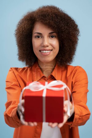 Foto de Mujer joven satisfecha sosteniendo caja de regalo envuelta en rojo, dando regalo, mirando sonriendo a la cámara, usando ropa de estilo casual. Estudio interior plano aislado sobre fondo azul - Imagen libre de derechos