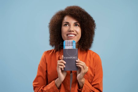 Téléchargez les photos : Portrait of happy curly haired woman holding passport and looking up with toothy smile, travelling. Indoor studio shot isolated on blue background - en image libre de droit