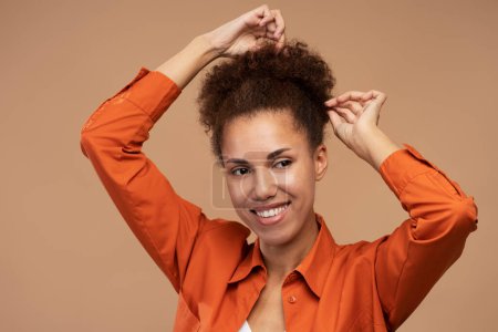 Foto de Portrait of cheerful curly african woman preparing hairstyle and looking away with toothy smile facial expression. Indoor studio shot isolated on brown background - Imagen libre de derechos