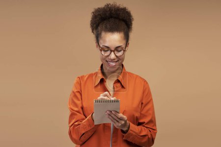 Foto de Portrait of smiling African American woman writing notes isolated on brown background. Student girl studying, education concept - Imagen libre de derechos