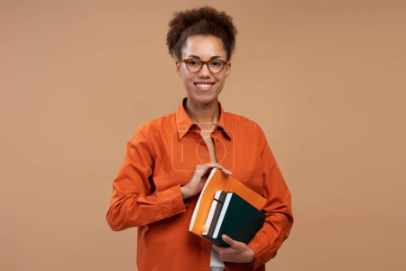 Foto de Portrait of smiling African American woman holding books isolated on brown background. Student studying looking at camera, education concept - Imagen libre de derechos
