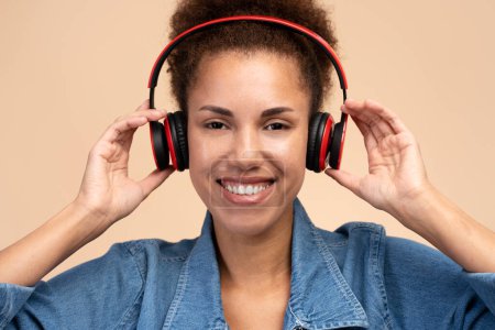 Téléchargez les photos : Happy funny woman standing and listening music with headphones, looking at camera with satisfied face, Indoor studio shot, isolated on beige background - en image libre de droit