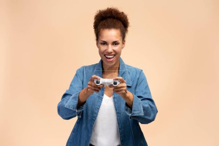 Téléchargez les photos : African American gamer woman jouer à un jeu vidéo en utilisant joystick sur fond beige isolé. Femme très heureuse regardant vers la caméra - en image libre de droit