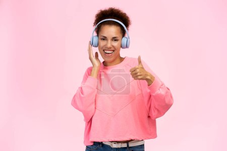 Foto de Happy funny woman standing and listening music with headphones, looking at camera with satisfied face. Indoor studio shot, isolated on pink background. Entertainment concept - Imagen libre de derechos