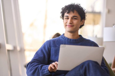 Téléchargez les photos : Portrait d'un beau sourire positif jeune homme latino-américain avec ordinateur portable, regardant avec confiance la caméra. Homme d'affaires télétravail dans un intérieur de bureau moderne. Les gens. Concept d'entreprise en ligne - en image libre de droit
