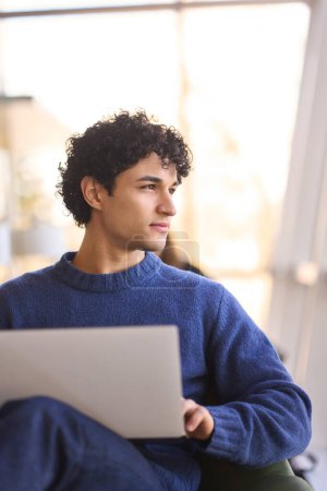 Photo for Close-up confident portrait of a handsome multitasking pensive Latin American man, looking aside while using laptop, telecommuting from a cozy modern office interior. People Business technology - Royalty Free Image