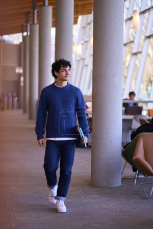 Photo for Handsome Hispanic male student, holding a laptop and notepad, walking on the hall of a modern library campus. Educated people and education concept - Royalty Free Image
