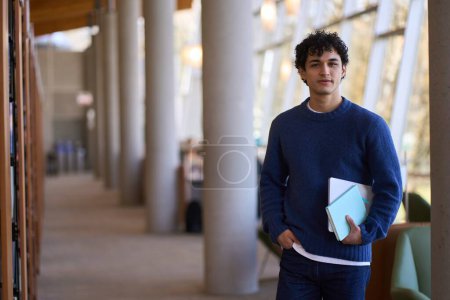 Téléchargez les photos : Portrait d'un jeune étudiant latino-américain, tenant un ordinateur portable et un bloc-notes, regardant avec confiance la caméra, debout sur le hall d'un campus de bibliothèque universitaire moderne. Éducation et concept des personnes - en image libre de droit