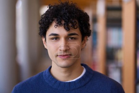 Portrait of handsome 20 years old young Latin American man, wearing blue knitted sweater, confidently looking at camera, standing against blurred background of bookshelves in a library or a bookstore