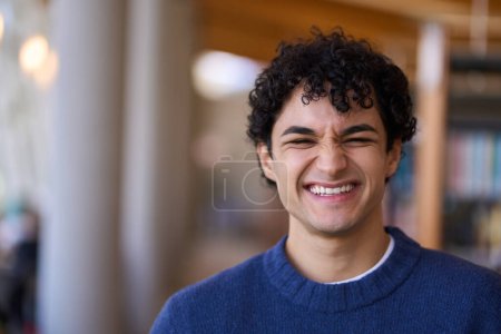 Photo for Handsome positive Latin American man, university student smiling a cheerful toothy smile looking at camera, standing against blurred background of bookshelves in a library or bookstore. People concept - Royalty Free Image