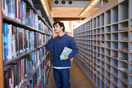 Photo for Pensive multi-ethnic male student, thoughtfully looking at bookshelves, searching books in the library campus of a high school or university institution. People. Erudition. Education. World Book Day - Royalty Free Image