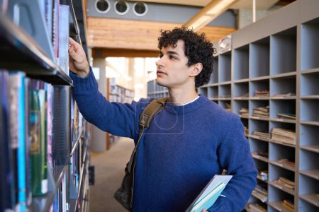 Photo for Handsome young dark-haired curly Latin American man in casual clothes with backpack, a university graduate student, selecting book from a bookshelf in the library campus. People and education concept - Royalty Free Image