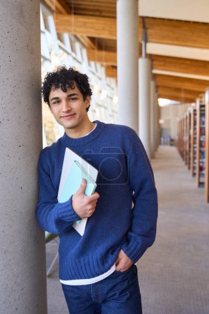 Photo for Confident Latin American young man, smart university graduate student in casual clothes, holding laptop and folder with docs, looking at camera while visiting the library campus. People and education - Royalty Free Image