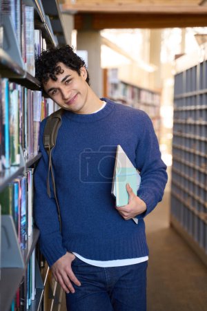 Photo for Handsome ethnic male student with backpack, cutely smiles, looking at camera while chooses books for his diploma project in the university library campus. People. Education. Knowledge. Learning - Royalty Free Image