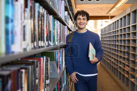 Photo for Handsome positive mixed race guy, a smart student standing by bookshelves in a library campus or bookstore, smiling a cheerful toothy smile looking at camera. Education Learning Erudite people concept - Royalty Free Image