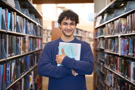 Foto de Retrato confiado de un guapo estudiante, chico latinoamericano, sonriendo mirando a la cámara, expresando emociones positivas, de pie junto a las estanterías con literatura en el campus de la biblioteca o librería - Imagen libre de derechos