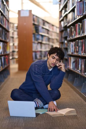 Photo for Hispanic multitasking smart male student talking on mobile phone, reading and searching information on a book while sitting at laptop on the floor, in the library campus.Education and people concept - Royalty Free Image