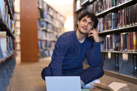 Téléchargez les photos : Pensif beau jeune homme latino-américain, un étudiant universitaire regardant attentivement et réfléchissant sur son projet de diplôme, assis sur le sol, en utilisant un ordinateur portable pour étudier sur le campus de la bibliothèque. Éducation - en image libre de droit