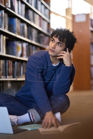 Photo for Handsome pensive Latin American young student talks on mobile phone, sitting on the floor in the library library campus and doing research for his diploma project, using printed literature and laptop - Royalty Free Image