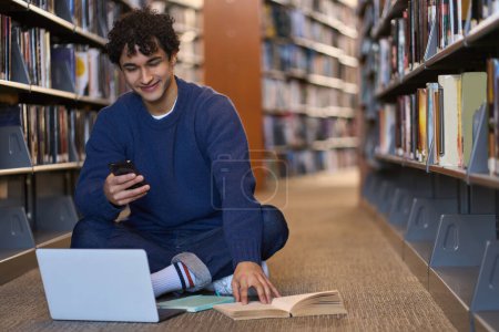 Photo for Handsome positive Latin American man, student spending time in library campus, using mobile phone and laptop, searching informations for his diploma or school project, sitting on floor by bookshelves - Royalty Free Image