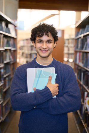 Photo for Smiling Hispanic young male student, holding copybook and laptop, looking at camera standing between bookshelves with literature and books in th campus. World Book Day. People and education concept - Royalty Free Image
