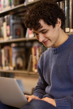 Photo for Young dark-haired curly Latin American guy, smart confident positive male student using laptop for study, sitting by shelves with literature and books in library campus. People. Technology. Education - Royalty Free Image