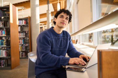 Foto de Estudiante masculino hispano joven pensativo usando el ordenador portátil, trabajando en un proyecto de diploma, mirando soñadoramente a un lado mientras busca información en Internet, navegando por sitios web en un campus moderno de biblioteca de innovación - Imagen libre de derechos