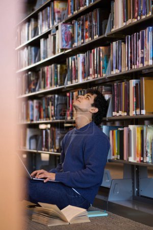 Téléchargez les photos : Multitâche jeune étudiant latino-américain coûteux, en utilisant un ordinateur portable pour étudier, assis sur le sol par des étagères avec de la littérature et des livres dans le campus de la bibliothèque universitaire. Les gens. La technologie. Éducation - en image libre de droit