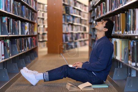 Photo for Pensive multitasking Hispanic young man, student leaning on bookshelf with literature, using laptop, writes text, makes researches while studies on his free time, sitting on floor in a library campus - Royalty Free Image