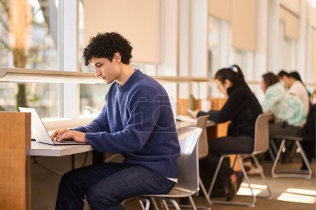 Foto de Multitarea, exceso de trabajo, seriedad Estudiante joven latinoamericano, en línea estudiando en un campus moderno de la biblioteca de innovación, sentado en un escritorio y escribiendo texto en el teclado del ordenador portátil, buscando información en la red - Imagen libre de derechos