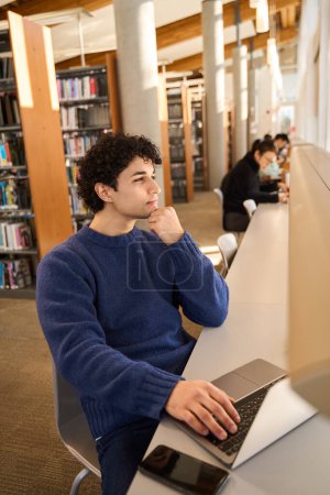 Foto de Estudiante masculino de raza mixta inteligente cuidadosamente mirando a un lado mientras trabaja en línea, estudiando en línea en un campus moderno de la biblioteca. Gente. Erudición. Educación. Enseñanza a distancia. Tecnología inalámbrica moderna - Imagen libre de derechos