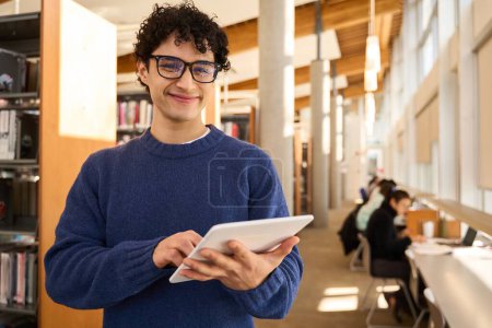 Photo for Positive smart multiethnic male student, wearing eyeglasses, holding a digital tablet and smiling, looking at camera, standing in the modern innovation library campus. People and education concept - Royalty Free Image