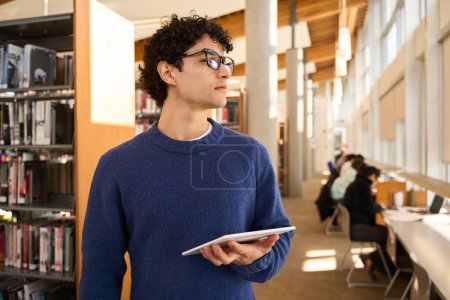 Photo for Confident smart Latin American young student, wearing eyeglasses, holding a digital tablet and thoughtfully looking aside, standing in a modern innovation library campus. People and education concept - Royalty Free Image