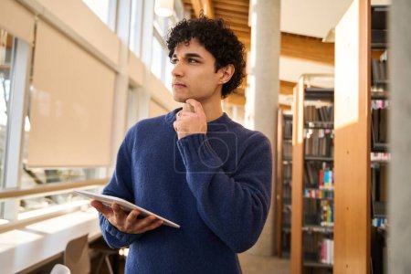 Photo for Pensive Latin American man, a smart university graduate student holding a digital tablet, working on his graduation project in a modern library campus. People. Erudition. Technology. Education - Royalty Free Image