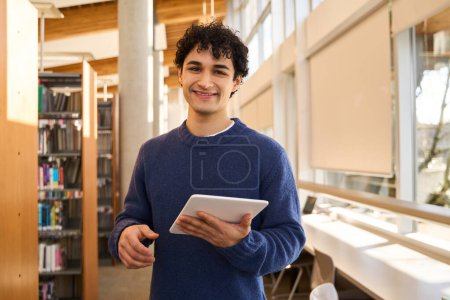 Photo for Handsome Latin American male student with digital tablet and smiling, looking at camera, visiting a modern innovation library campus, making researches for his diplome project. People and education - Royalty Free Image