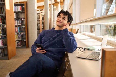 Photo for Positive smiling Hispanic man, dreamily looking aside, sitting at desk with laptop and holding a mobile phone in his hands. Male student in the library campus. People. Education. Learning concept - Royalty Free Image