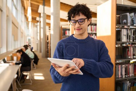 Photo for Handsome positive Latin American male student wearing eyeglasses, using a digital tablet, browsing internet resources, making researches for his diploma project, working online in the library campus - Royalty Free Image