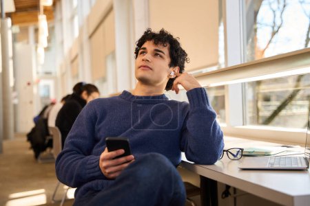 Photo for Pensive serious Latin American male student holding smartphone, listening to an online lecture on earphones, sitting at desk with laptop in a modern library campus. Smart guy during exam preparations - Royalty Free Image